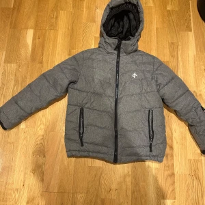 Grå pufferjacka från Cross Sportswear - Säljer en grå pufferjacka från Cross Sportswear med huva och dragkedja. Jackan har långa ärmar och praktiska fickor med dragkedjor. Perfekt för kyliga dagar med sin vadderade design.