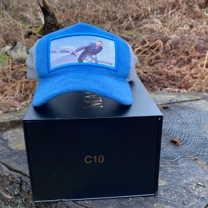 Blå keps med nätpaneler - Snygg blå keps med grå nätpaneler och justerbart spänne bak. Framsidan pryds av en tryckt bild. Perfekt för en avslappnad stil.