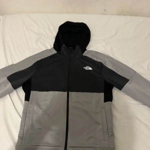 The North Face kofta  - Säljer en snygg grå och svart kofta från The North Face med huva och dragkedja. Perfekt för kyligare dagar. Koftan har långa ärmar och praktiska fickor framtill.