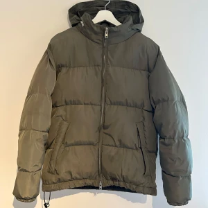 Olivgrön pufferjacka Brixtol Textiles - Säljer en olivgrön pufferjacka med avtagbar huva och dragkedja framtill. Jackan har långa ärmar och elastisk nederkant för extra komfort. Perfekt för kyliga dagar. Använd men i gott skick