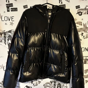 Svart pufferjacka med huva - Snygg svart pufferjacka från Zara med huva och dragkedja. Jackan har en glansig yta och två fickor framtill. Perfekt för kyliga dagar. Använd 1 gång.