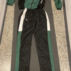 Grön och svart träningsjacka från Lacoste - Säljer en grön och svart träningsjacka från Lacoste med vita detaljer. Jackan har en dragkedja framtill och långa ärmar. Perfekt för sportiga aktiviteter eller en avslappnad stil.man får med hela dressen för ett rimligt pris