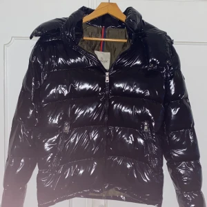Svart glansig pufferjacka från Moncler - Säljer en svart glansig pufferjacka från Moncler. Jackan har en dragkedja framtill och två fickor med dragkedjor. 