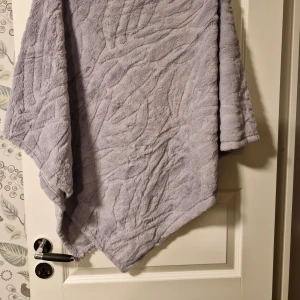 Ljusgrå fleeceponcho - Mysig ljusgrå poncho i fleece som är perfekt för kyliga dagar. Den har en lös passform och är enkel att slänga över axlarna för extra värme. Perfekt för att mysa hemma eller som ett extra lager ute.