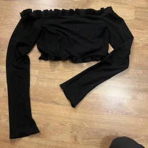 Svart ribbad offshoulder topp från NLY One - Snygg svart ribbad offshoulder topp från NLY One med volangdetaljer vid hals och nederkant. Perfekt för en trendig look. Passar bra till både jeans och kjol.