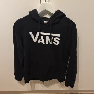 Svart hoodie från Vans - Säljer en svart hoodie från Vans med klassisk logga i vitt på framsidan. Tröjan har en bekväm huva med dragsko och en praktisk magficka. Perfekt för en avslappnad stil.