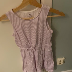 Ljuslila randig klänning från H&M - Säljer en söt ljuslila och vit randig klänning från H&M. Klänningen är ärmlös med en elastisk midja som ger en bekväm passform. Perfekt för varma dagar!