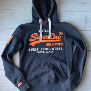 Superdry hoodie - Säljer en svart hoodie från Superdry med tryck i orange och vitt. Den har en klassisk känguruficka och justerbar huva med vita snören. Perfekt för en avslappnad stil. Storlek XS. 