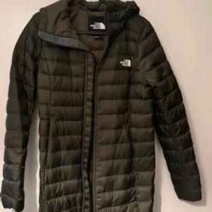 Olivgrön dunjacka från The North Face - Säljer en olivgrön dunjacka från The North Face. Jackan har en lång design med dragkedja och huva, perfekt för kyliga dagar. Den är quiltad för extra värme och komfort. En stilren och funktionell jacka för alla tillfällen.