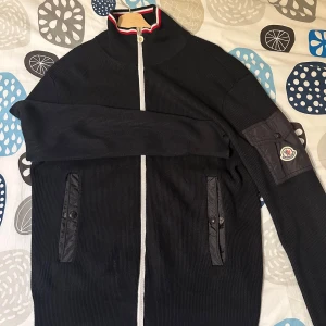 Svart stickad jacka från Moncler - Säljer en svart stickad jacka från Moncler med dragkedja och ribbade detaljer. Jackan har en broderad logotyp på ärmen och en randig krage i rött och vitt. Perfekt för kyligare dagar!