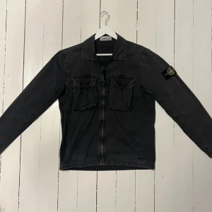 Svart overshirt från Stone Island - Snygg svart overshirt från Stone Island med dragkedja och två bröstfickor. Den har en klassisk krage och långärmade ärmar. Märkets ikoniska patch syns på ärmen. Perfekt för en stilren look.