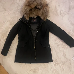 Svart Woolrich parkajacka med pälsfodrad huva - Säljer en stilren svart parkajacka från Superdry med en mysig pälsfodrad huva. Jackan har dragkedja och knappar framtill samt praktiska fickor. Perfekt för kyliga dagar. Köparen står för frakt.