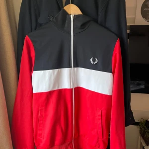 Röd,vit och svart tröja från Fred Perry - Säljer en snygg röd och svart fullzip från Fred Perry med vit rand över bröstet. Tröjan har en dragkedja framtill och det klassiska Fred Perry-emblemet på bröstet. Som ni ser på tredje bilden finns det två fläckar på de vita, vad jag märkt syns dessa främst i ett viss ljus. Medans till vanligt vardags ljus är de inte lika lätt synligt. Storlek M i bra skick 