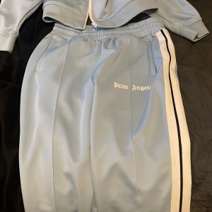 Palm Angels Tracksuit - Säljer detta baby blåa Palm Angels dress i mycket bra skick! Finns små saker man kan behöva sy, ett litet slit vid fickorna på byxorna som inte syns utan har du en förälder eller nära som kan sy kan de få de att se helt nya ut! Bältet tillkommer vid köp av denna dress! Byxorna storlek L, och tröjan storlek M.