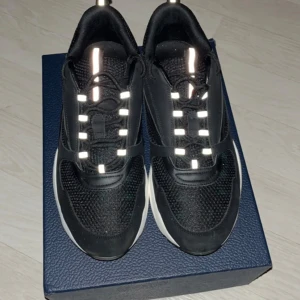 Dior B22 - Snygga svarta sneakers från Dior med vit sula och reflekterande detaljer på snörningen. Skorna har en modern design med en kombination av syntetmaterial och textil för en stilren look.