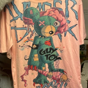 Rosa t-shirt med grafiskt tryck - Cool rosa t-shirt med ett stort grafiskt tryck av en tecknad figur i grönt och rosa. Texten 'Gum Toy' och andra färgglada detaljer ger en unik look. Perfekt för dig som gillar streetwear och vill sticka ut med en färgglad stil.