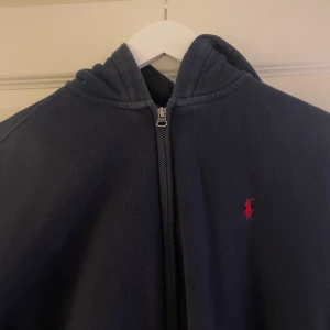 Mörkblå hoodie från Ralph Lauren - Säljer en snygg mörkblå hoodie från Ralph Lauren med dragkedja och en liten röd logga på bröstet. Perfekt för en avslappnad stil. Hoodien har en klassisk design med huva och är i storlek S(barn storlek XL).