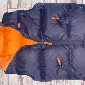 Mörkblå dunväst från Napapijri - Säljer en snygg mörkblå dunväst från Napapijri med orange foder. Västen har dragkedja och två praktiska fickor med orange detaljer. Perfekt för kyliga dagar!skriv vid intresse och frågor