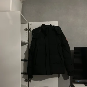 Canada Goose Wyndham  - Säljer en svart dunjacka från Canada Goose med huva och broderad logotyp på ärmen. Jackan har en pufferdesign med långa ärmar och dragkedja framtill. Perfekt för kalla vinterdagar.men där det står M i naken har lossnat men det e M 