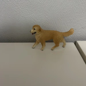 Schleich hund  - En liten figur av en hund i plast, med ljusbrun färg och detaljerad pälsstruktur. Perfekt som dekoration eller leksak.