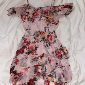 Blommig rosa klänning med volanger - Charmig rosa klänning med blommigt mönster och volanger. Den har smala axelband och en offshoulder-design.