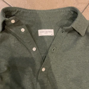 Grön pikétröja från John Henric - Säljer en stilren grön pikétröja från John Henric. Tröjan har långa ärmar och knappar framtill. Perfekt för en avslappnad men ändå uppklädd look. Tröjan är i toppskick och passar er som är 176-182
