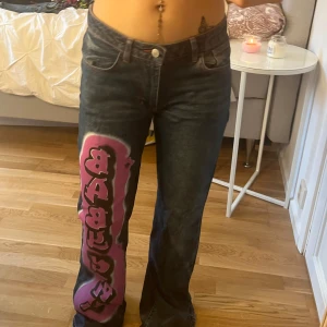 Blåa low rise jeans med rosa detaljer - Coola blåa jeans med rosa text 'BABY GIRL' längs benet och ett hjärta broderat på bakfickan. De har en bootcut-stil och är lågmidjade, vilket ger en trendig look. Perfekta för att sticka ut med sina unika detaljer. De har rosa sömmar vilket ser väldigt snyggt ut.