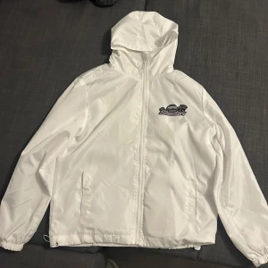 Vit vindjacka med huva - Säljer en vit vindjacka från Trapstar med huva och dragkedja. Jackan har ett tryck med texten 'London Shooters' på bröstet och ryggen. Perfekt för blåsiga dagar.