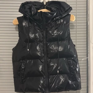 Svart dunväst från Shein - Snygg svart dunväst från Shein med glansig yta och dragkedja framtill. Västen har en huva med dragsko och är perfekt för kyligare dagar. Den är både stilren och praktisk.