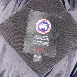 Wyndhamn Parka Canada Goose - Säljer en svart dunjacka från Canada Goose med huva och flera praktiska fickor. Jackan har både dragkedja och knappar framtill samt en broderad logotyp på ärmen. Perfekt för kalla vinterdagar. En knapp borta annars slitren använd bara 1 vinter. Black Kabel Wyndham Parka modell.