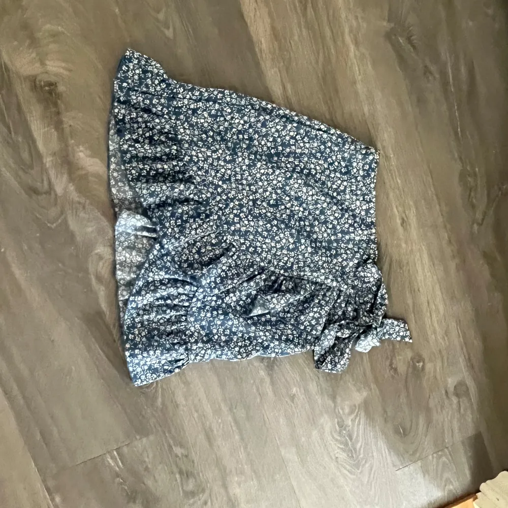Söt blommig omlottkjol i blått med volangdetaljer. Perfekt för en somrig look. Kjolen har en knytning i sidan som ger en fin passform.. Hameet.