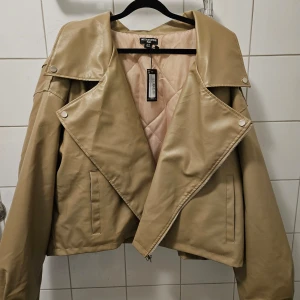 Beige skinnjacka från prettylittlewoman - Snygg beige skinnjacka från prettylittlewoman med dragkedja och knappar. Jackan har en cool asymmetrisk design och långa ärmar. Perfekt för att ge din outfit en edgy touch. Den är aldrig använd, storlek 54