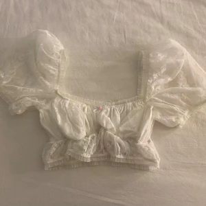 Vit spetsblus från For Love & Lemons - Säljer en romantisk vit spetsblus från Victoria’s Secret. Blusen har puffiga korta ärmar och en söt knytning framtill. Perfekt för sommarens alla tillfällen. 🌸