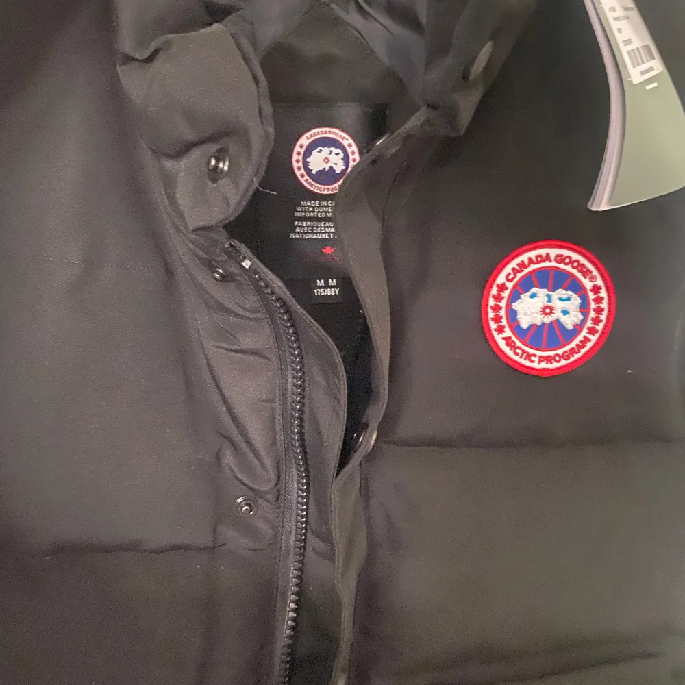 Säljer en svart dunväst från Canada Goose nysick Västen har en dragkedja och knappar framtill samt två praktiska fickor. Perfekt för kyliga höst- och vinterdagar. Den har en klassisk stil med det ikoniska märket på bröstet. Passar både till vardags och mer äventyrliga aktiviteter.. Toppar.