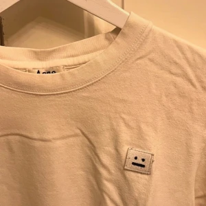 Vit t-shirt från Acne Studios - Säljer en stilren vit t-shirt från Acne Studios i bomull. Den har en liten broderad logga på bröstet som ger en cool touch. Perfekt för vardagsbruk eller att styla upp med en jacka. Skön och enkel att matcha med det mesta!