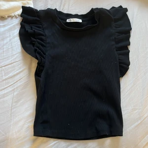Svart ribbad topp från Zara - Snygg svart ribbad topp från Zara med volangdetaljer på ärmarna. Perfekt för både vardag och fest! Toppen är i ett mjukt och stretchigt material som sitter bekvämt.