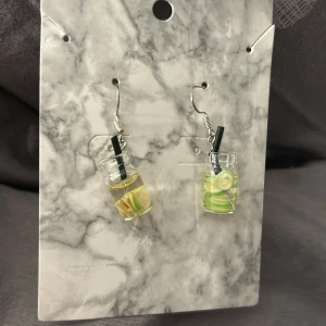Örhängen med drinkmotiv - Unika örhängen med små glas som ser ut som drinkar, dekorerade med lime och sugrör. Perfekta för att ge en rolig och somrig touch till din outfit. nickelfria och med pluppar
