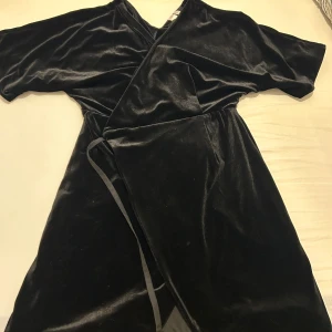 Svart omlottklänning i sammet - Elegant svart omlottklänning i mjuk sammet från Mango. Klänningen har korta ärmar och ett knytband i midjan för en smickrande passform. Perfekt för en stilren look.