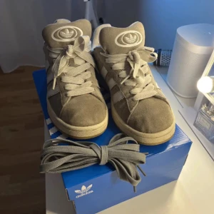 Grå Adidas Campus sneakers - Säljer ett par grå Adidas Campus sneakers i mocka med vita detaljer och snörning. Skorna är i bra skick. Har dock tappat bort dom gråa skosnörena dom kommer med. Perfekta för en avslappnad stil och passar till både vardag och fest.