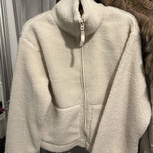 Vit teddyjacka med dragkedja - Mysig vit teddyjacka med dragkedja framtill och hög krage. Perfekt för kyliga dagar med sin fluffiga och varma design. Jackan har en avslappnad passform och två praktiska fickor framtill.