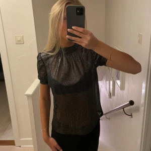 Svart glittrig blus - Snygg svart glittrig blus med korta ärmar. Perfekt för festliga tillfällen! Blusen har en rund hals och ett elegant fall som ger en stilren look.