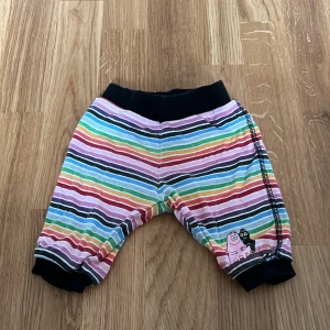 Randiga byxor från H&M - Färgglada randiga byxor från H&M med svart resår i midjan och vid bensluten. De har ett sött tryck av en rosa figur på framsidan. Perfekta för en lekfull stil!