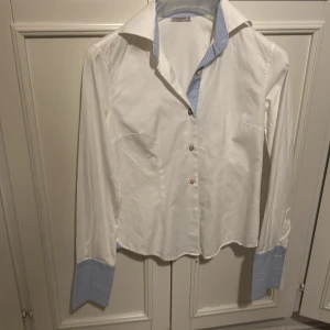 Vit skjorta från Forque - Snygg vit skjorta från Forque med blå randiga detaljer på krage och ärmslut. Tillverkad i 100% bomull för en skön och luftig känsla. Perfekt för en stilren look.