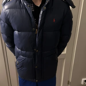 Blå dunjacka från Polo Ralph Lauren - Säljer en snygg blå dunjacka från Polo Ralph Lauren i storlek S den passar även xs i barn storlek. Jackan har en dragkedja och knappar framtill, samt en praktisk huva. Perfekt för kalla vinterdagar. Den är i mycket bra skick och håller dig varm och stilren. Passar både till vardags och lite finare tillfällen.