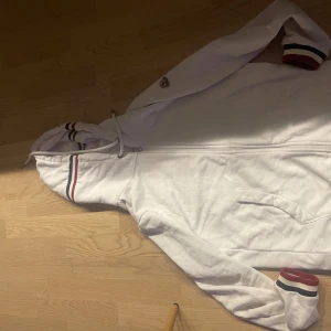 Vit huvtröja från Moncler - Säljer en snygg vit huvtröja från Moncler i mycket bra skick. Den har en dragkedja framtill och ribbade muddar med ränder i blått och rött. Perfekt för höst och vår, med en stilren design och Moncler-logga på ärmen. Passar både till vardags och lite finare tillfällen.