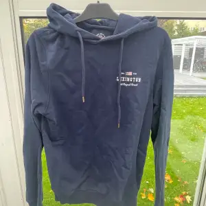 Säljer en snygg mörkblå hoodie från Lexington i bra skick. Den har en klassisk design med dragsko i huvan och en liten logga på bröstet. Perfekt för höst och vår när du vill vara både bekväm och stilren. Passar till både jeans och joggers! Ser ut som färgskiftningar men det är bara på bilden. 