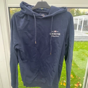 Mörkblå hoodie från Lexington - Säljer en snygg mörkblå hoodie från Lexington i bra skick. Den har en klassisk design med dragsko i huvan och en liten logga på bröstet. Perfekt för höst och vår när du vill vara både bekväm och stilren. Passar till både jeans och joggers! Ser ut som färgskiftningar men det är bara på bilden. 