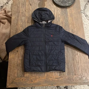 Svart och grå pufferjacka från Ralph Lauren - Säljer en stilren pufferjacka från Ralph Lauren i både svart och grå färg. Jackan har en broderad logotyp på bröstet, dragkedja och huva. När man vänder ut och in på den så byter den färg. Perfekt för kyligare dagar.