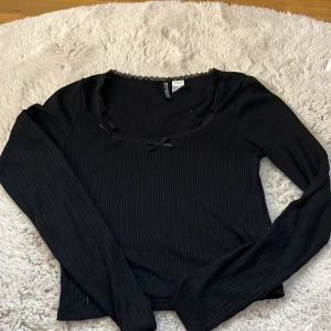 Svart ribbad topp från Divided - Säljer en svart ribbad topp från Divided i storlek S. Den har långa ärmar och en söt spetsdetalj runt halsringningen med en liten rosett framtill. Perfekt för både vardag och fest! 🖤