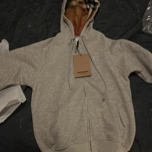 Grå hoodie från Burberry - Snygg grå hoodie från Burberry med klassiskt rutigt mönster i huvan. Tröjan har dragkedja och justerbara snören vid huvan. Perfekt för en stilren och bekväm look. Pris ej hugget i sten